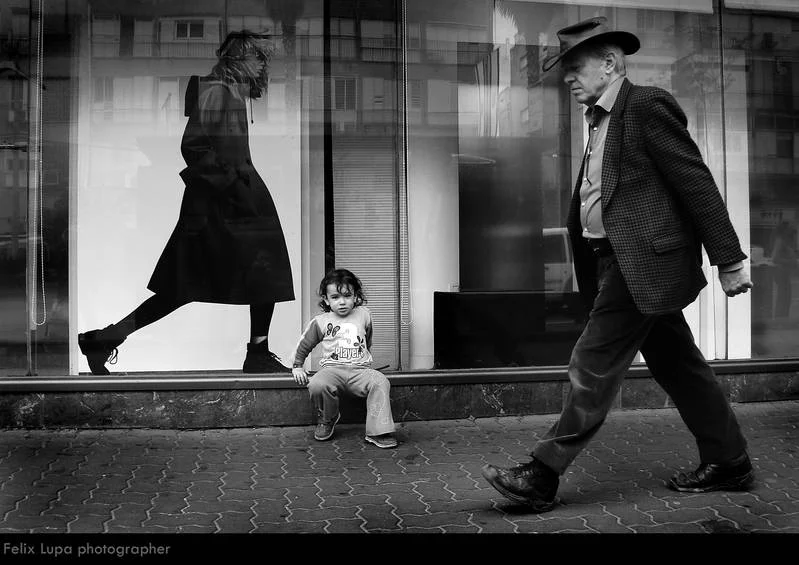 a child sitting infront of a shop while a man passes by the street